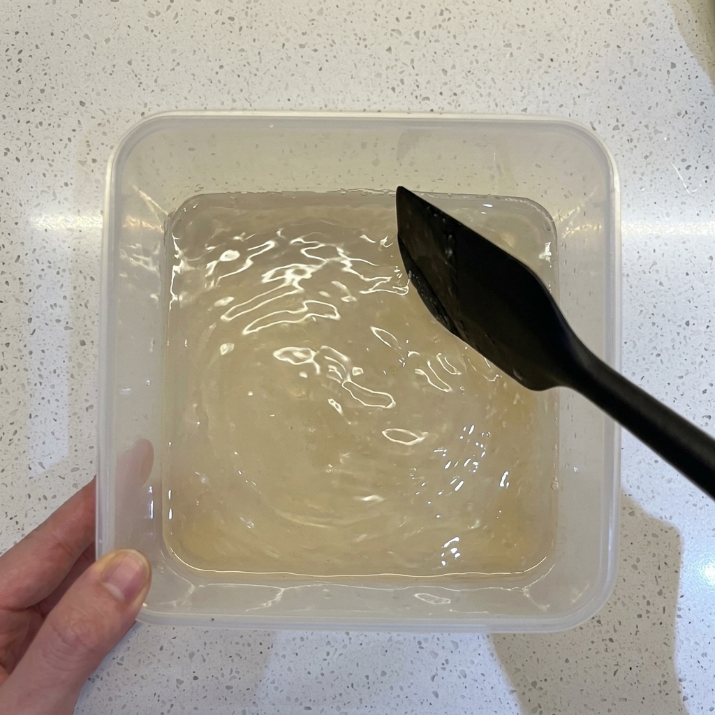 Una espátula negra mezclando harina, agua y levadura en un recipiente plástico transparente.