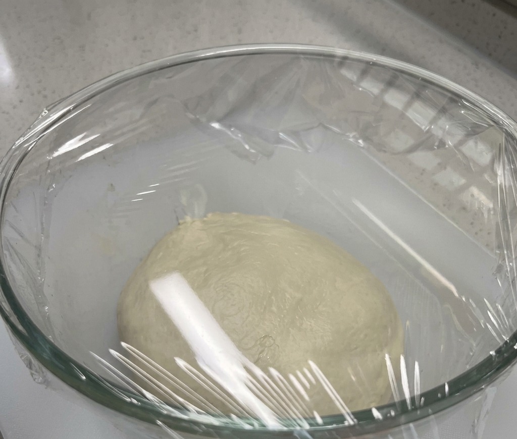 Una bola de masa de pizza lisa en el fondo de un bol de cristal sellado con film plástico.