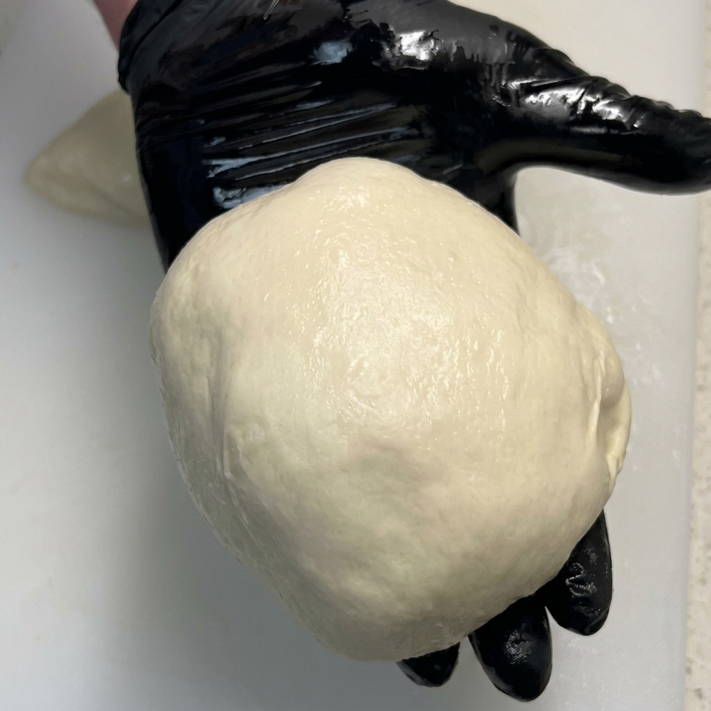 Una bola de masa de pizza perfectamente lisa y redonda en la palma de una mano con guante negro.