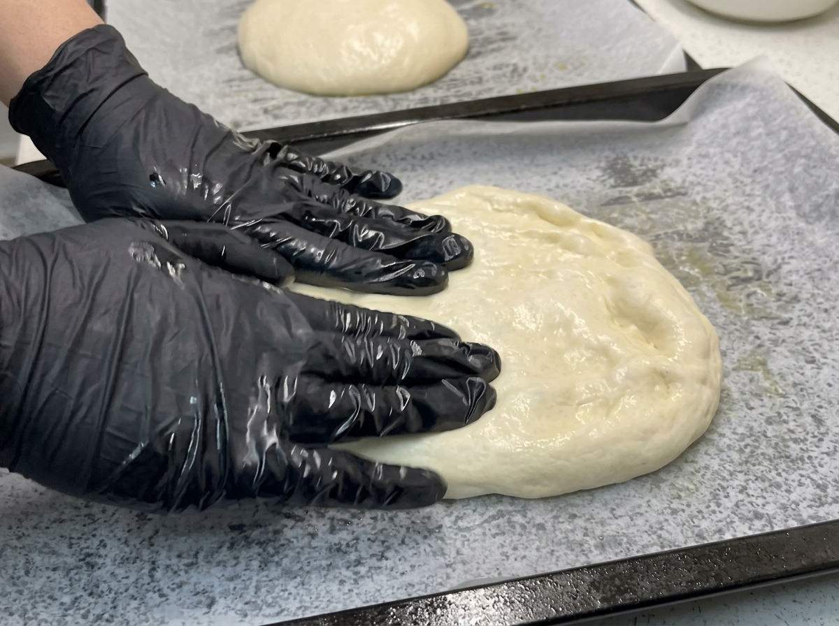 Manos con guantes presionando y estirando una masa de pizza suave sobre una bandeja con papel de horno.