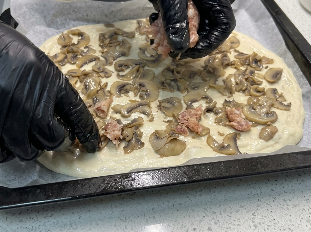 Manos colocando trozos de salchicha cruda sobre una masa de pizza que ya tiene los champiñones salteados.