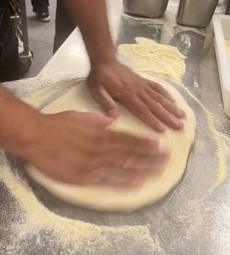 Manos de un chef estirando una masa de pizza hasta formar un círculo plano en un mostrador de acero.