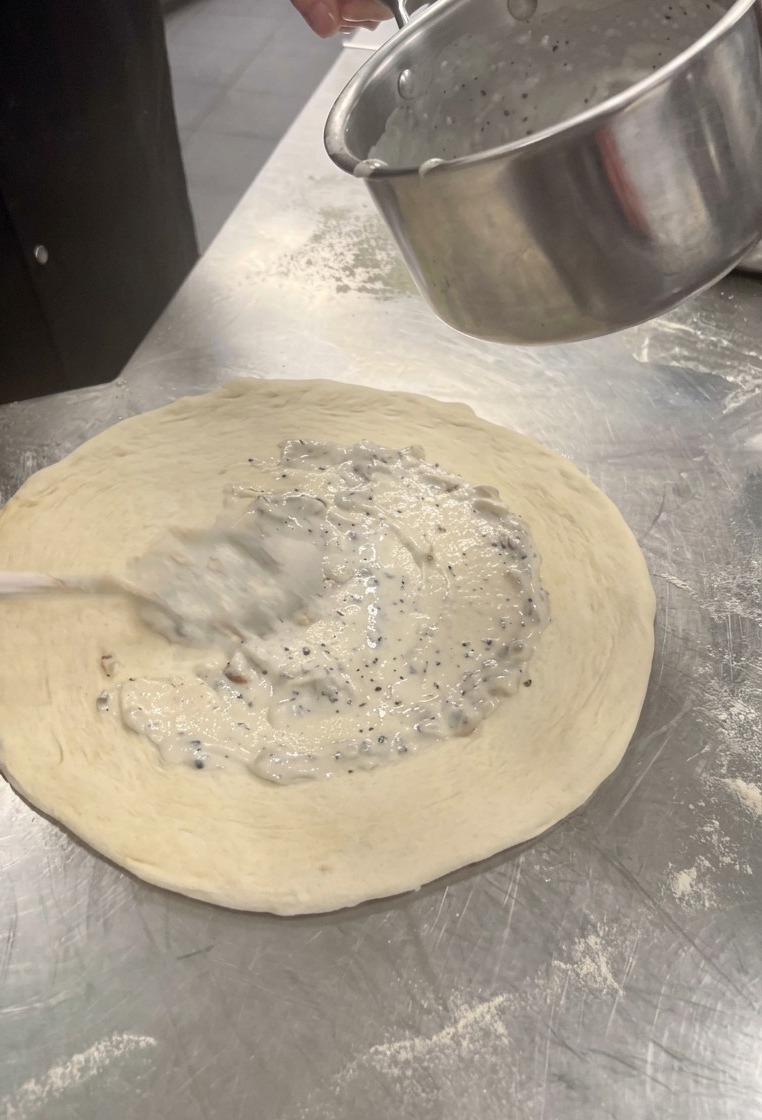 Extendiendo salsa cremosa de champiñones sobre el centro de la masa de pizza con una cuchara.