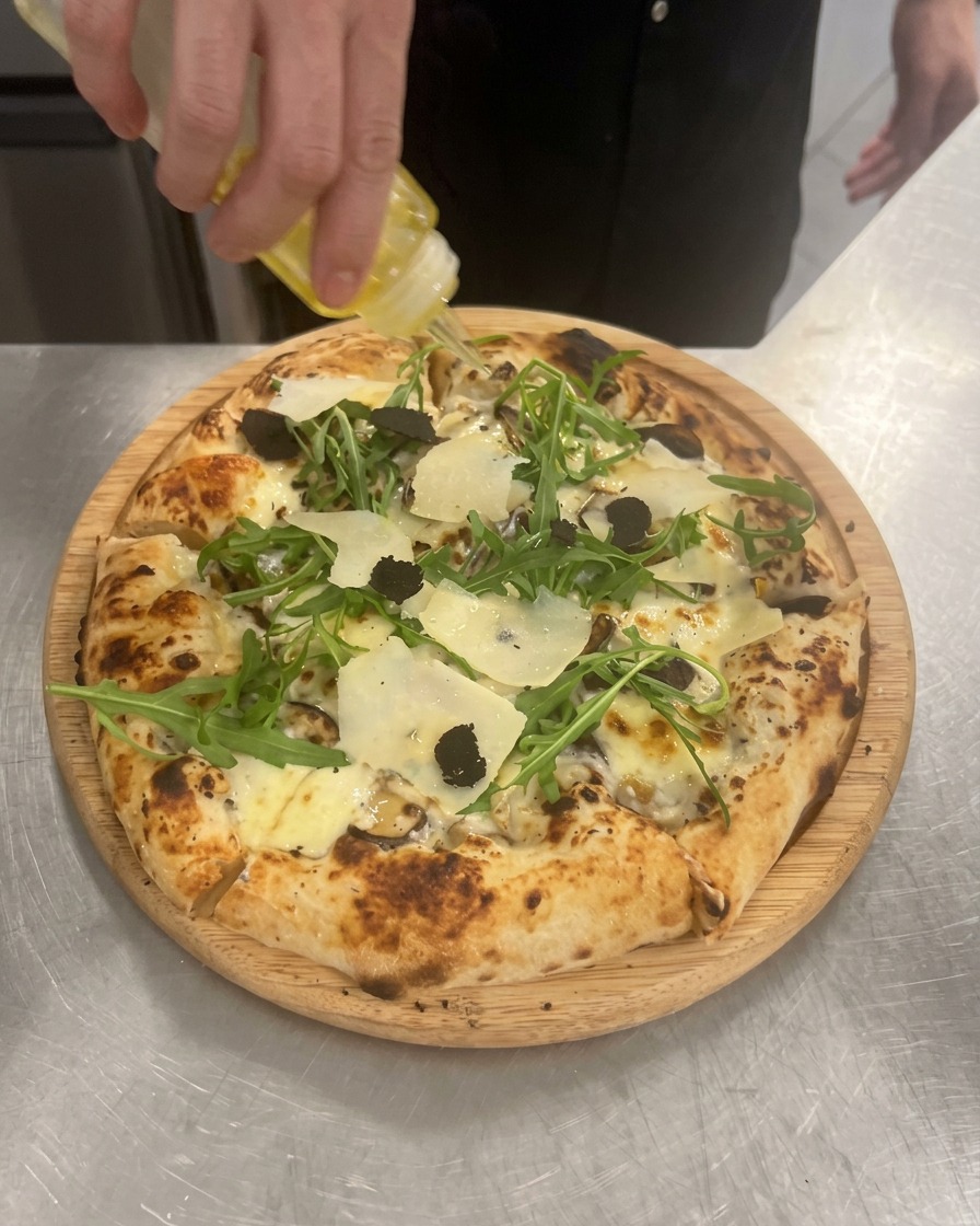 Mano vertiendo un hilo de aceite de trufa sobre una pizza terminada de champiñones, rúcula y trufa.