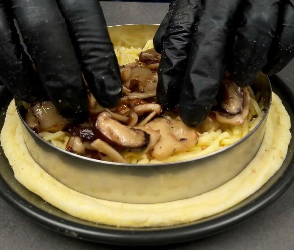 Manos con guantes negros colocando setas variadas cocinadas sobre una capa de queso mozzarella en la base de la pizza.