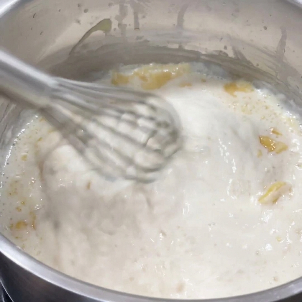 Un batidor removiendo una salsa blanca espesa y burbujeante en una olla.