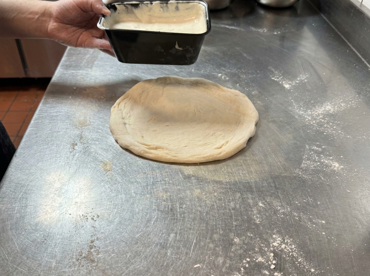 Salsa blanca vertiéndose sobre una masa de pizza cruda en una mesa de acero.