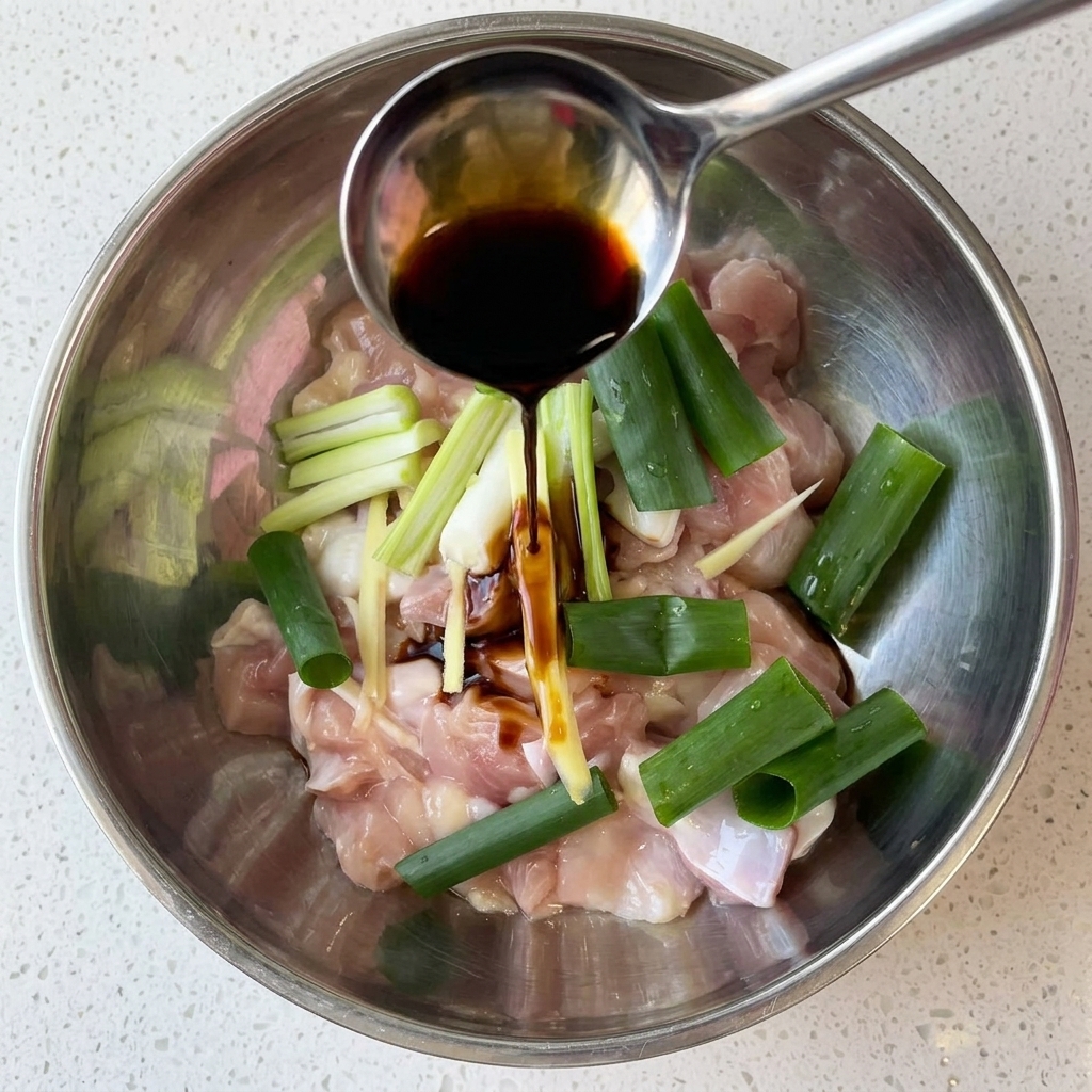 Trozos de pollo crudo en un bol metálico marinándose con salsa de soja oscura, cebolleta y jengibre.