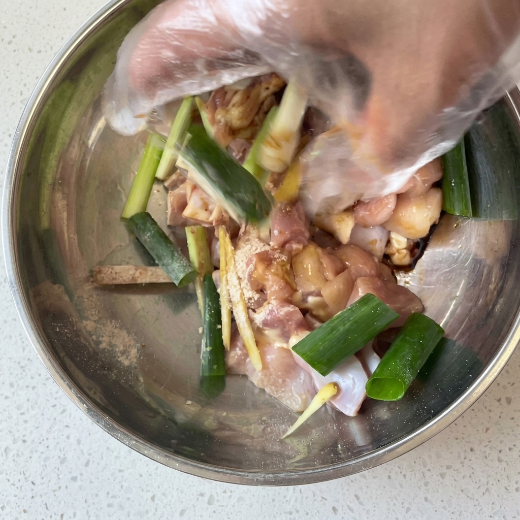 Una mano con guante mezclando trozos de pollo crudo con cebolleta, jengibre y marinada en un bol de metal.