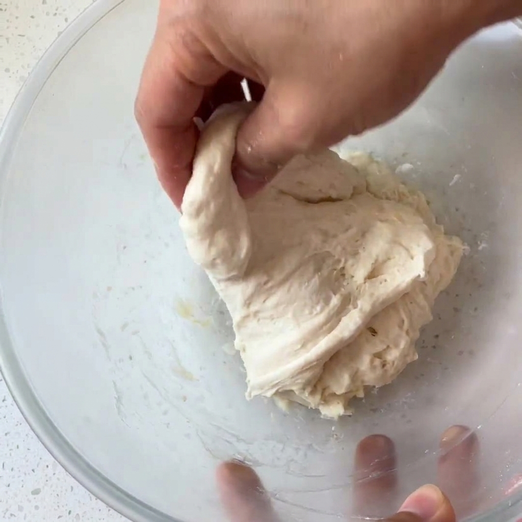 Mano agarrando y plegando una bola de masa húmeda y pegajosa dentro de un bol de cristal.