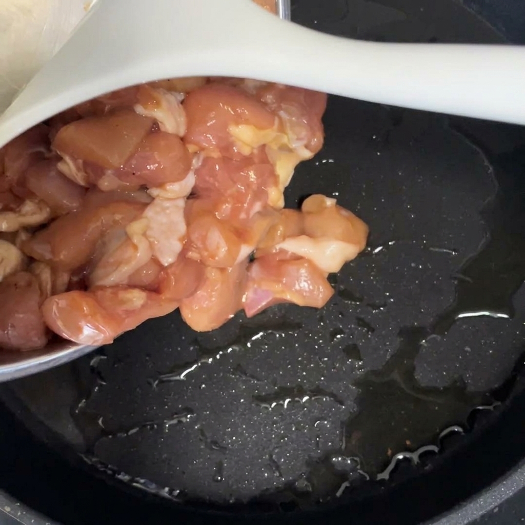 Trozos de pollo marinado crudo vertiéndose en una sartén negra con aceite caliente.