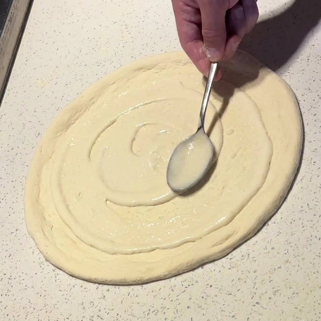Una cuchara de metal extendiendo salsa blanca cremosa en movimientos circulares sobre la base de pizza.
