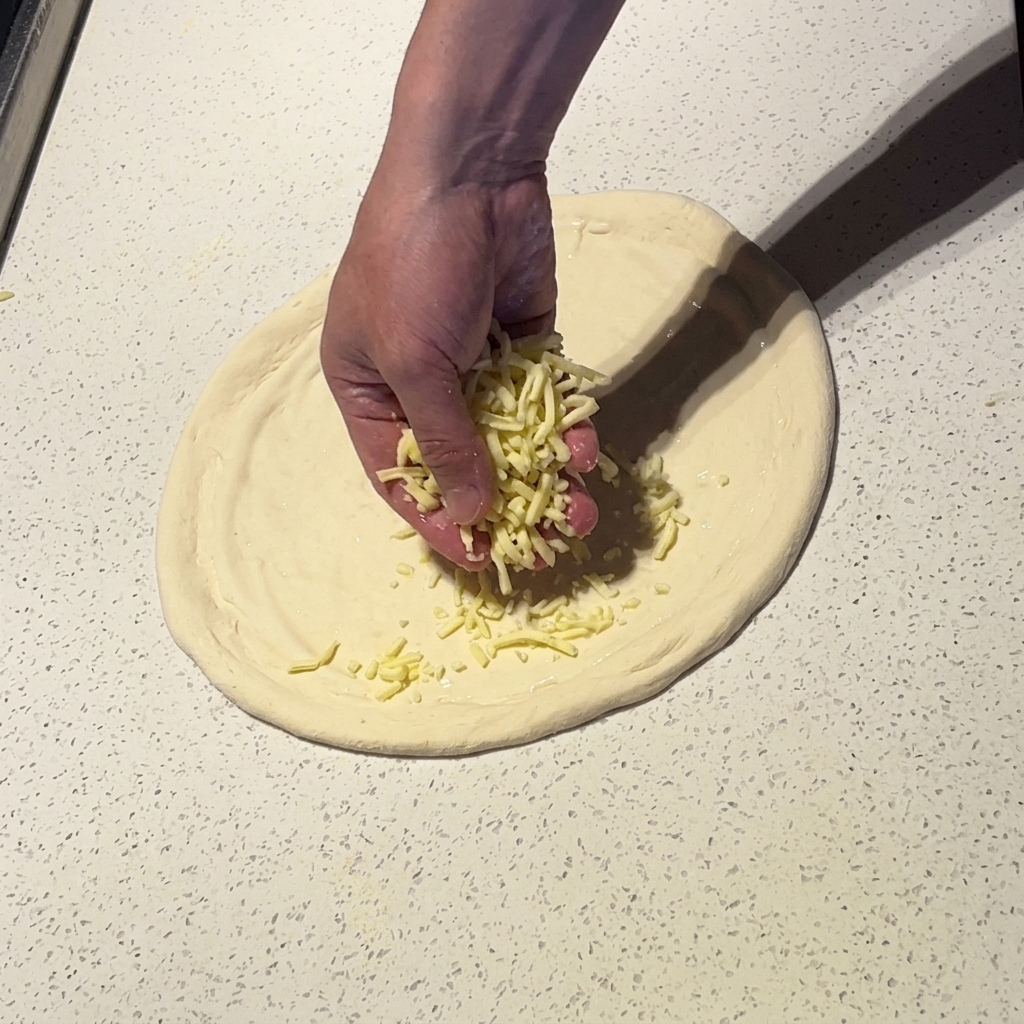 Una mano espolvoreando queso mozzarella rallado sobre la base de pizza con salsa blanca.