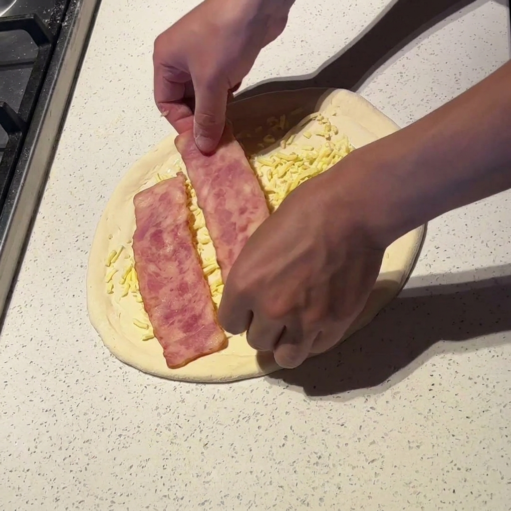 Manos colocando tiras anchas de bacon ligeramente cocido sobre la pizza con queso.