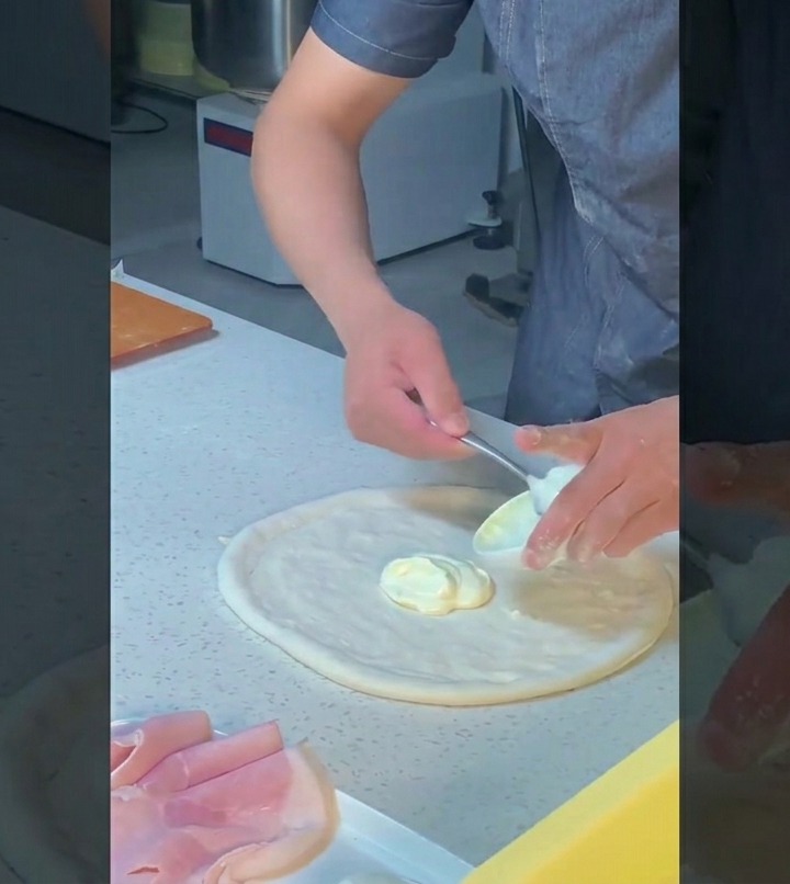 Cocinero usando el dorso de una cuchara para extender salsa blanca sobre la masa con movimientos circulares.