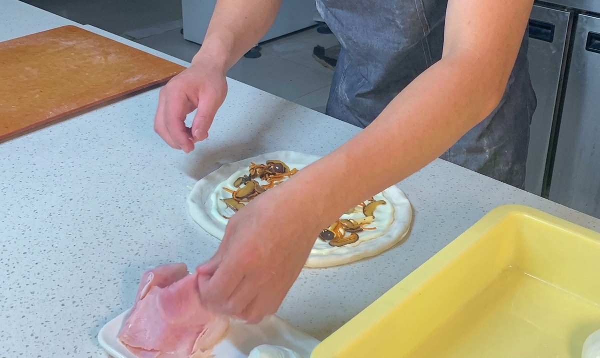 Manos troceando láminas de jamón y repartiéndolas sobre la pizza.