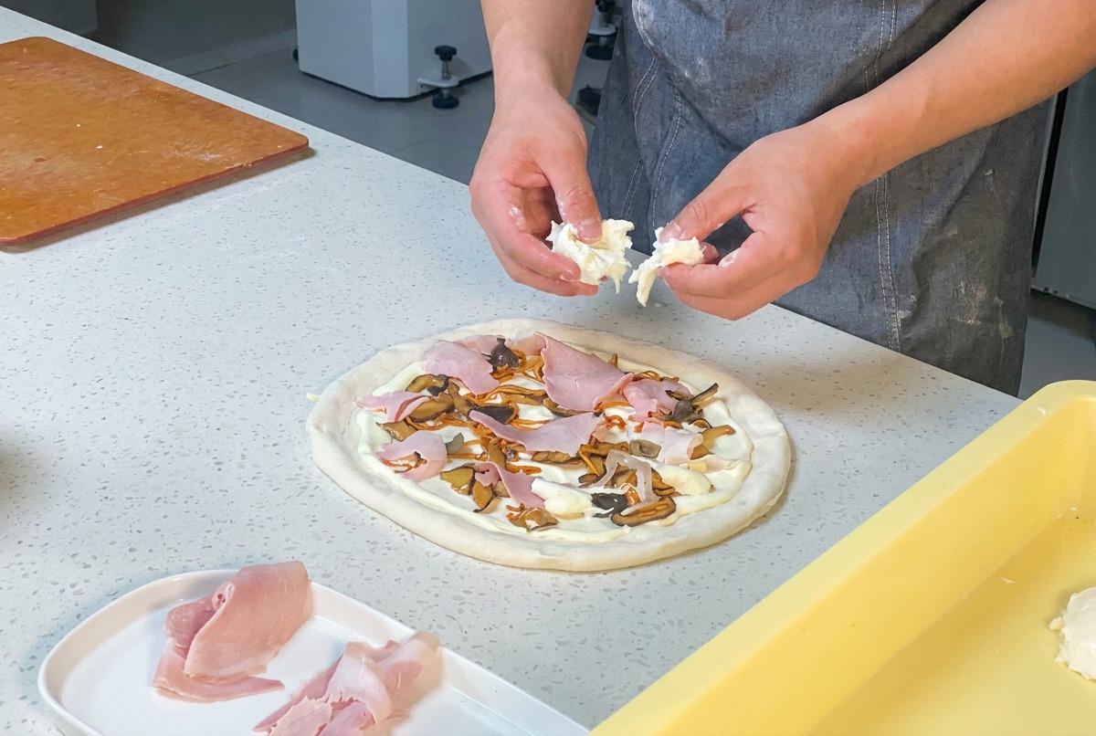 Manos desmenuzando mozzarella fresca sobre la pizza de jamón y champiñones.