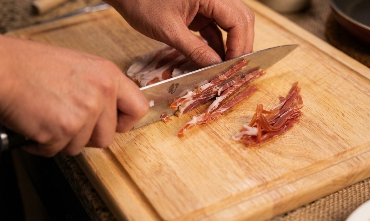Manos usando un cuchillo de chef para cortar tiras finas de jamón curado en una tabla de madera.