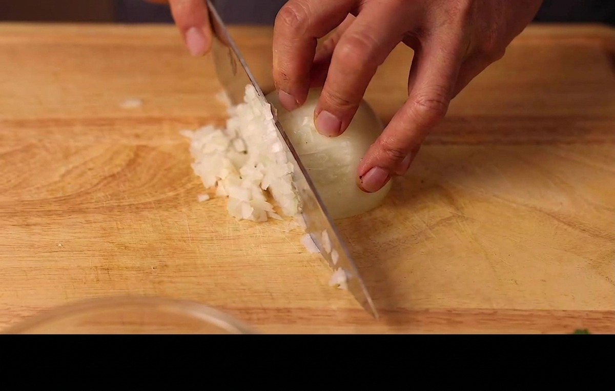 Manos picando finamente media cebolla blanca en una tabla de cortar de madera.