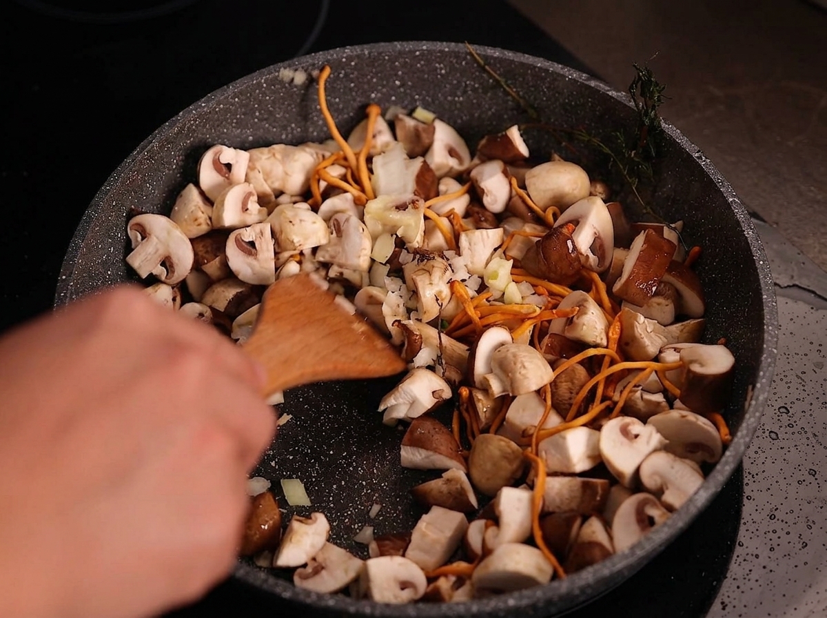 Una espátula de madera mezclando cebolla picada y ajo laminado en una sartén con setas cocinadas.