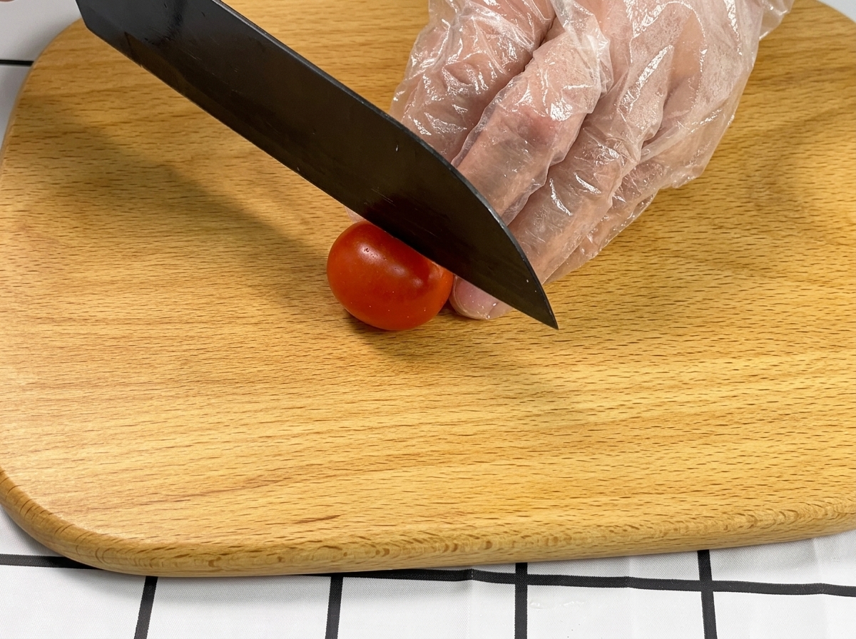 Mano con guante sujetando un tomate cherry mientras un cuchillo negro lo corta por la mitad sobre una tabla de madera.