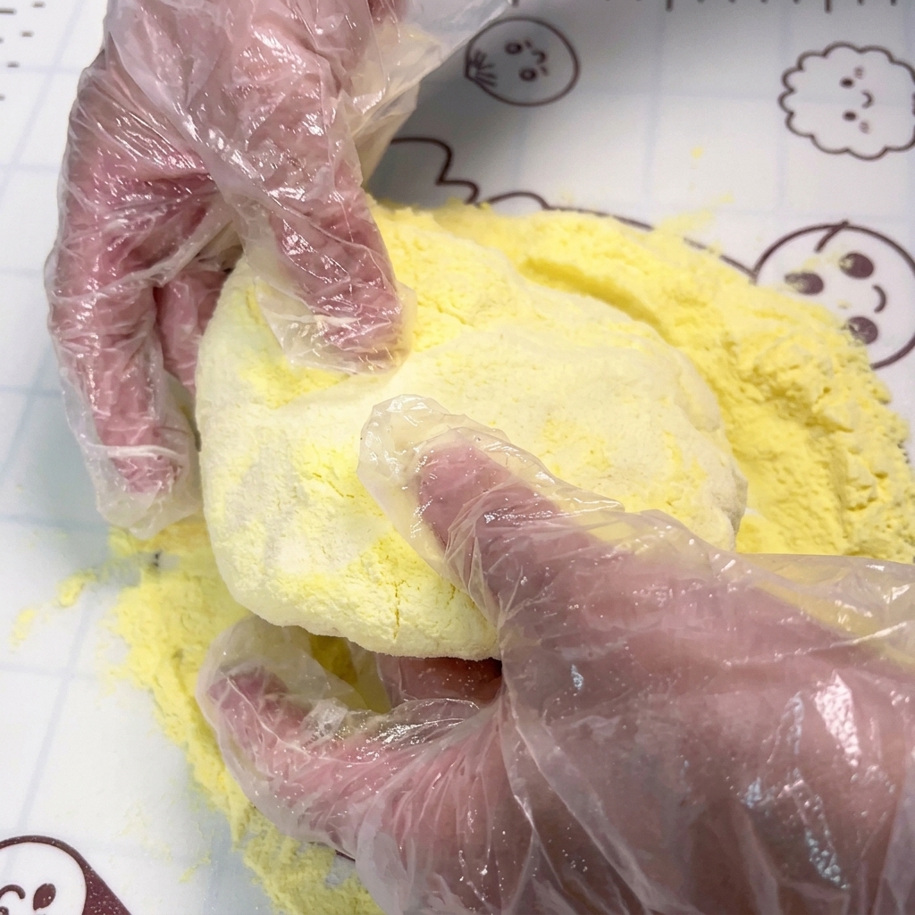Manos con guantes presionando y estirando una pieza redonda de masa sobre una superficie cubierta de harina de maíz amarilla.