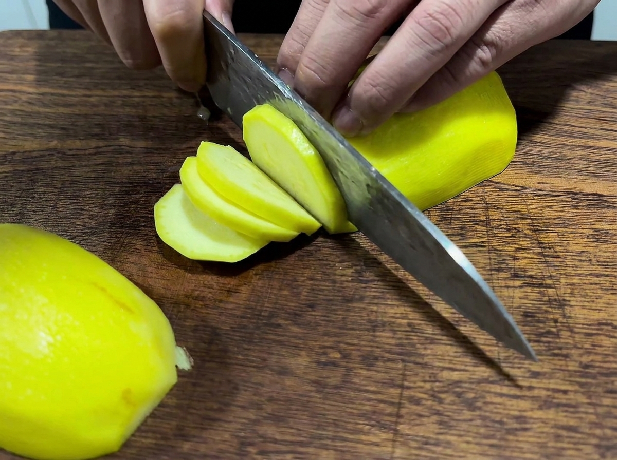 Manos usando un cuchillo grande para cortar una papa amarilla pelada sobre una tabla de madera.