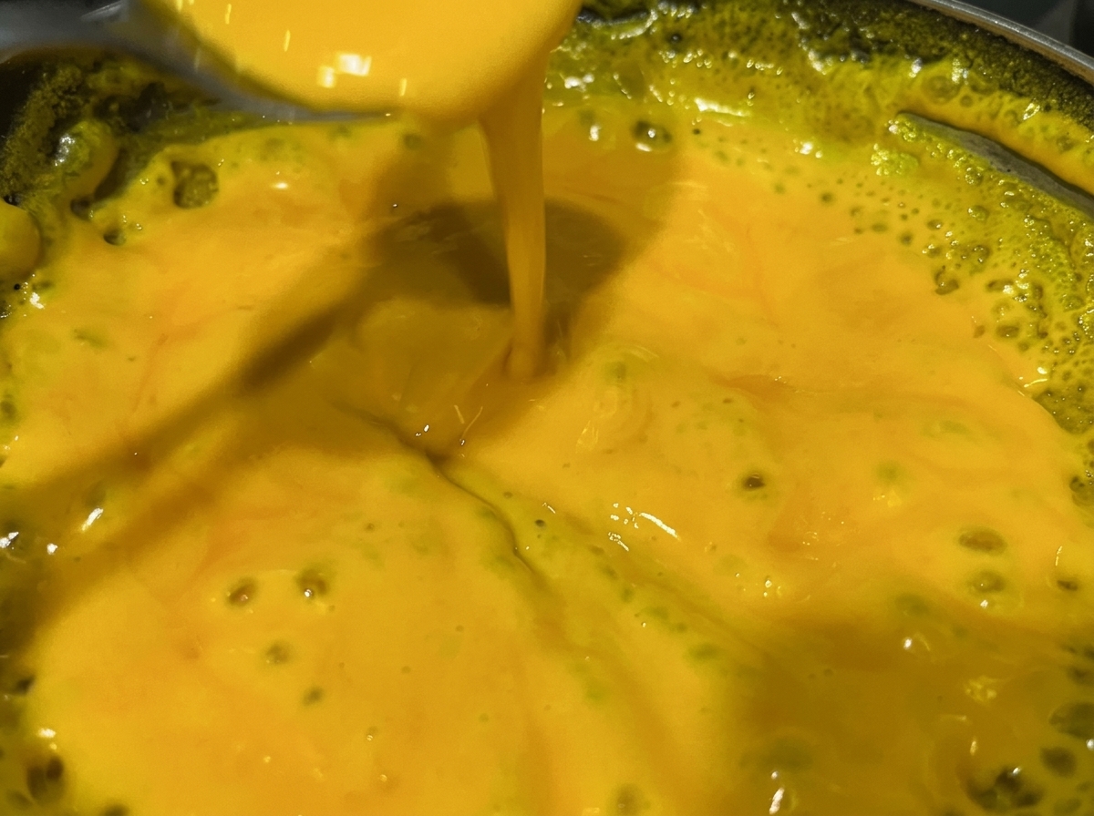 Una cuchara removiendo una salsa de queso amarilla brillante en una sartén metálica.