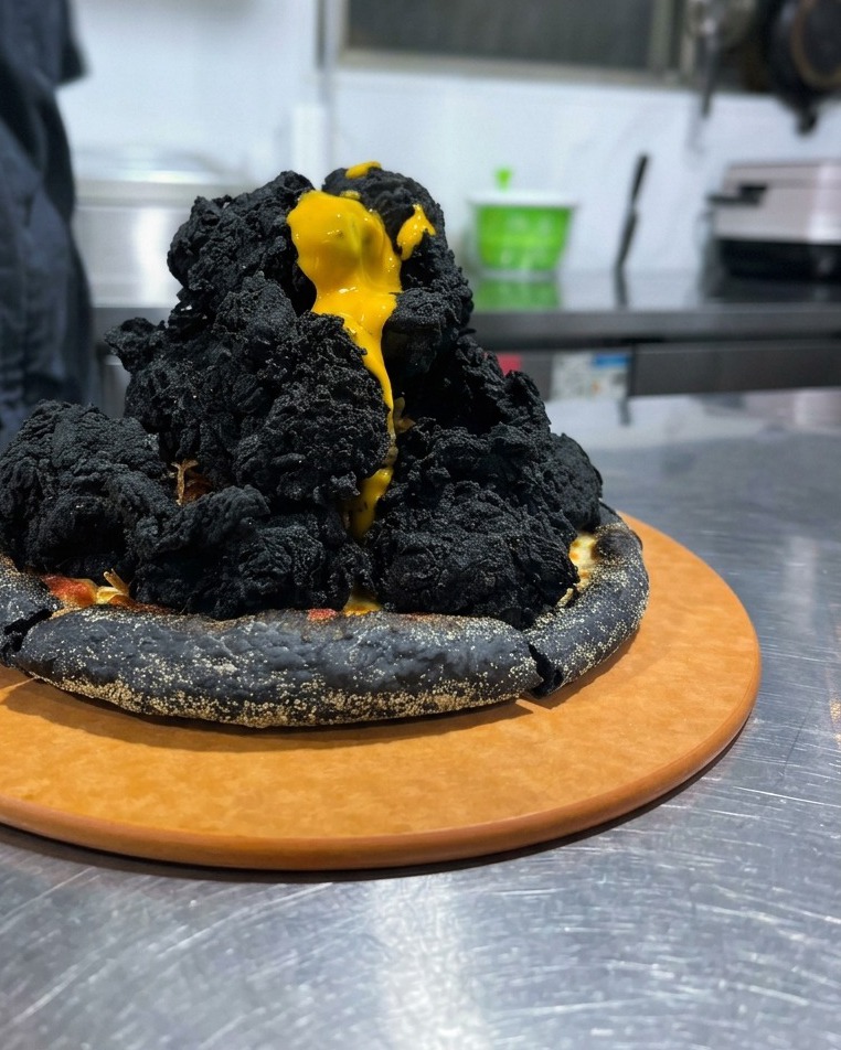Salsa de queso amarilla brillante goteando como lava sobre una montaña de pollo frito negro en una pizza negra.
