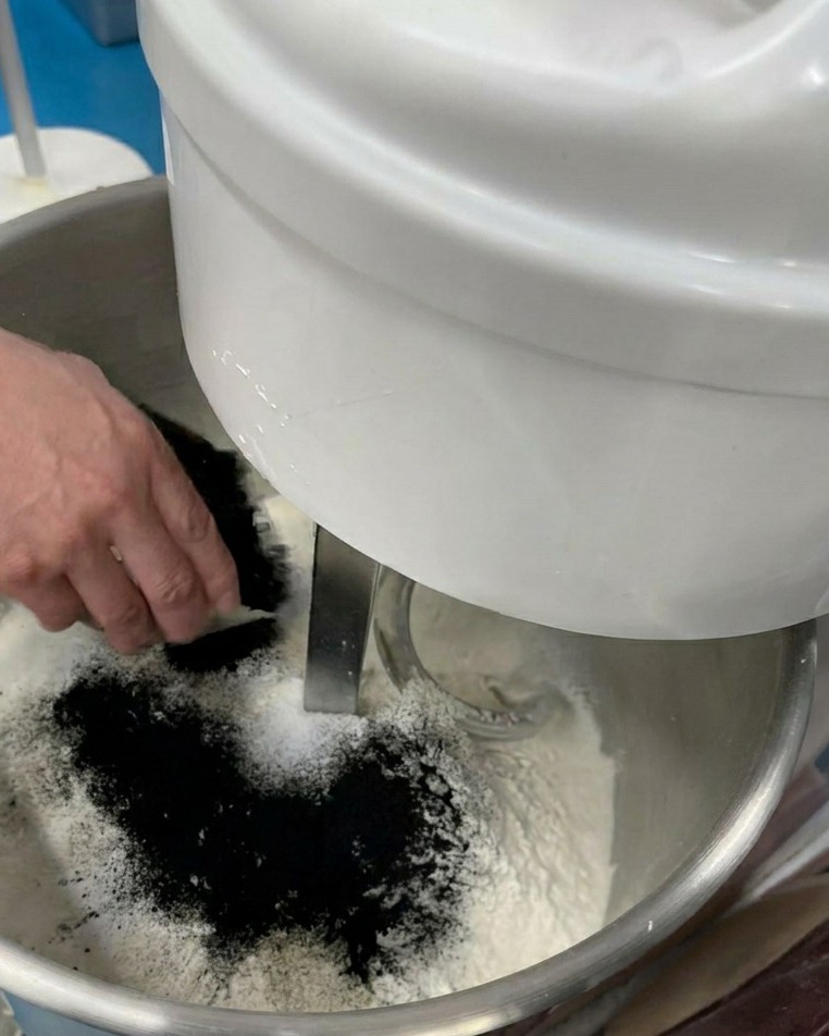 Una mano vertiendo un polvo negro fino en un bol de mezcla lleno de harina y otros ingredientes secos.