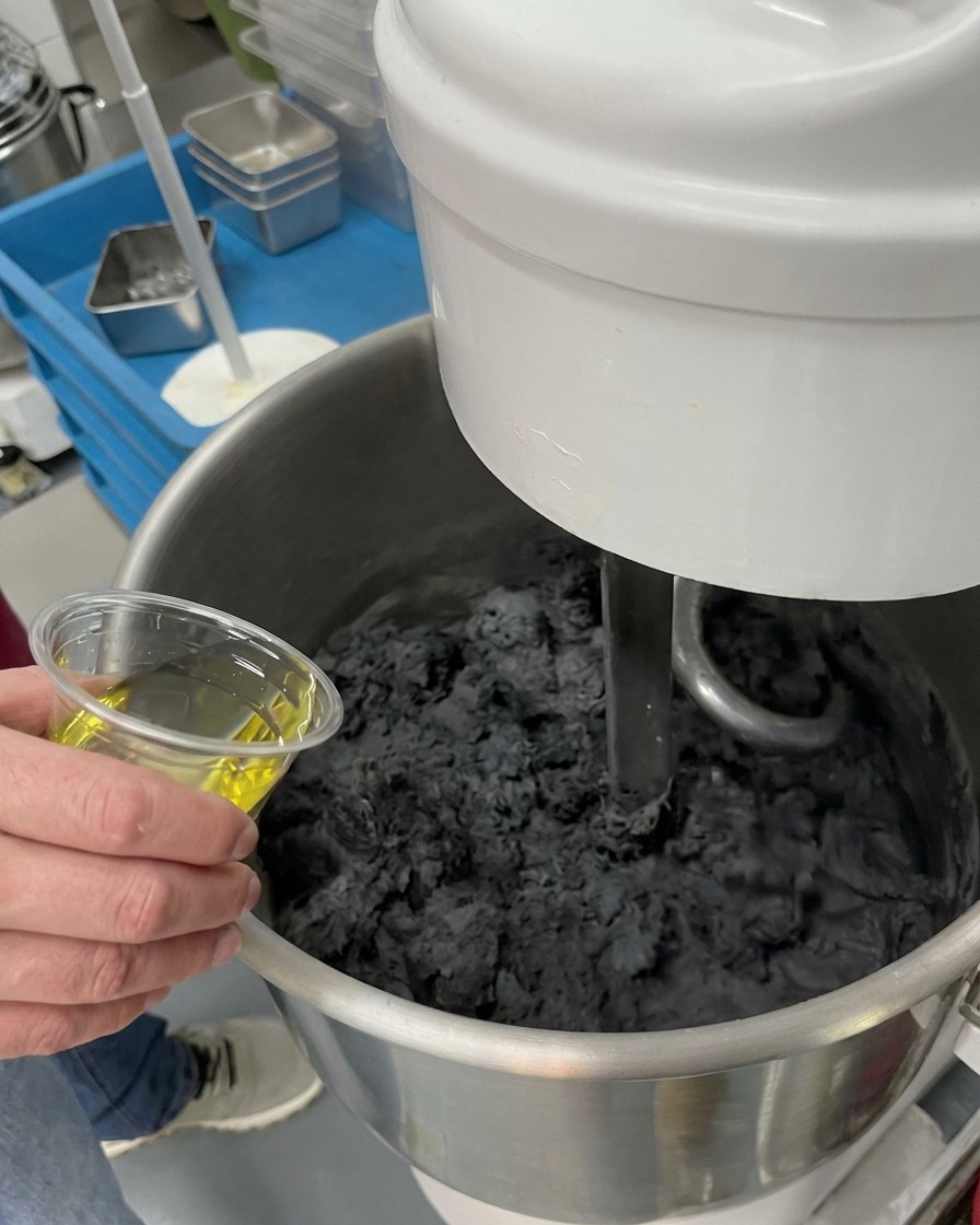 Una mano vertiendo aceite de cocina amarillo desde una taza transparente en un bol con masa negra espesa.