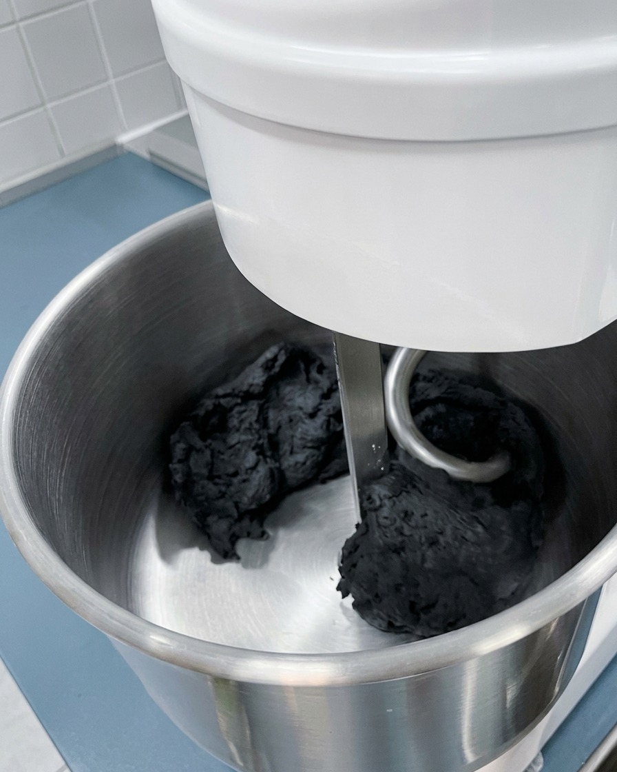 Primer plano del gancho de una amasadora trabajando una gran masa de color negro intenso en un bol de acero inoxidable.