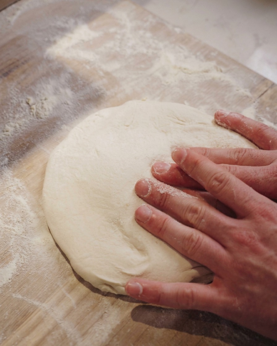 Dos manos presionando suavemente el centro de una masa de pizza sobre una superficie enharinada.