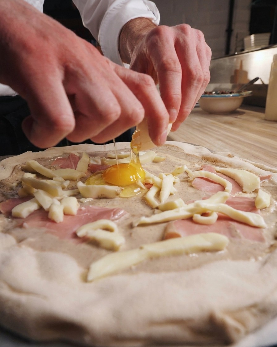 Manos de chef cascando un huevo en el centro de la pizza preparada.