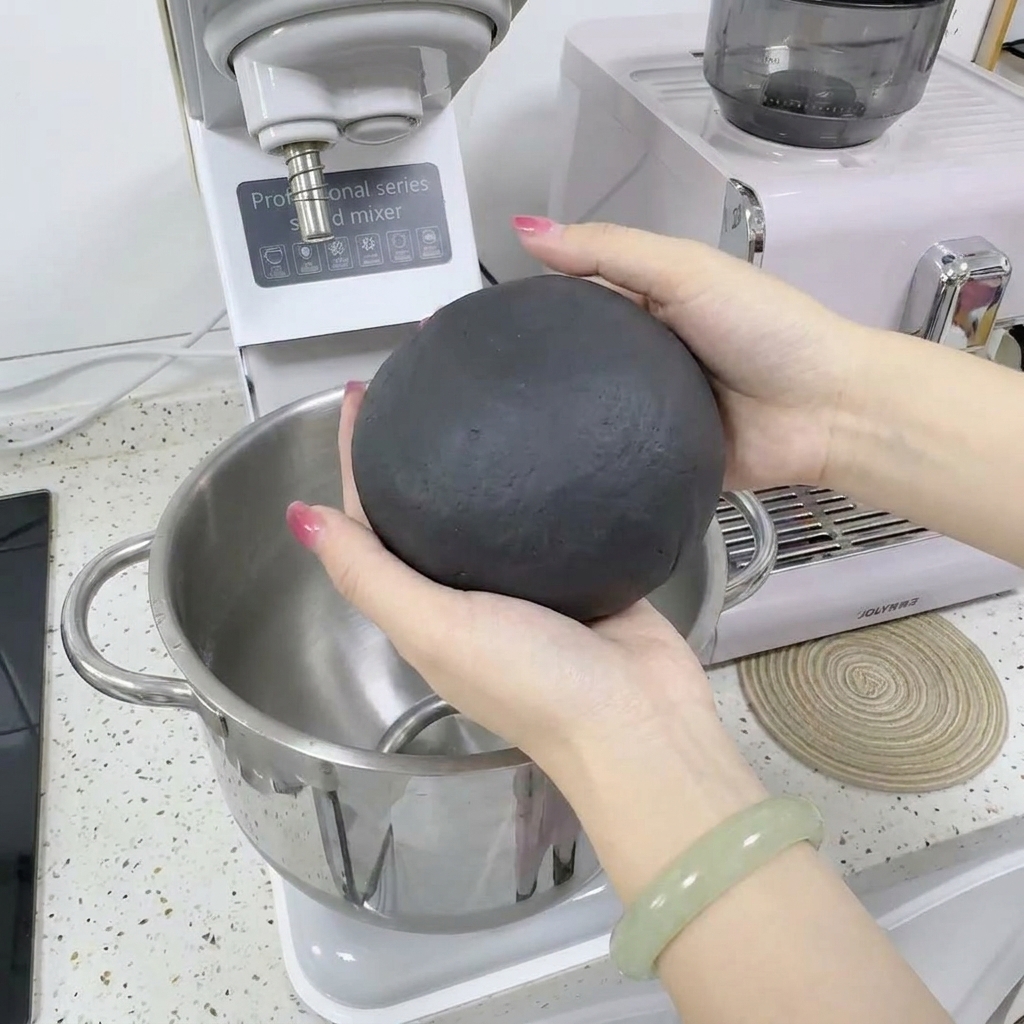 Dos manos sosteniendo y dando forma a una bola de masa gris oscuro frente a una batidora blanca.