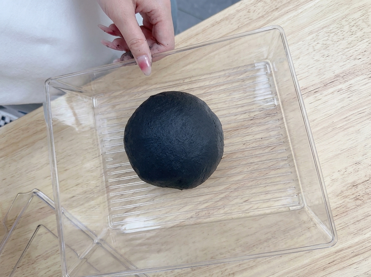 Una mano colocando la bola de masa negra en un recipiente de plástico transparente sobre una mesa de madera.