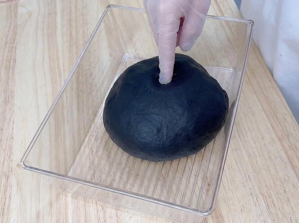 Un dedo con guante presionando la masa negra fermentada en un recipiente de plástico transparente.