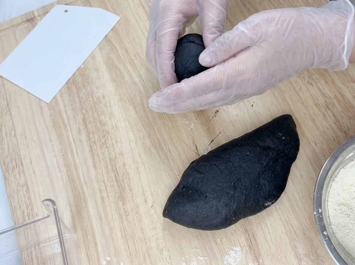 Manos con guantes rodando una porción de masa negra sobre una superficie de madera.