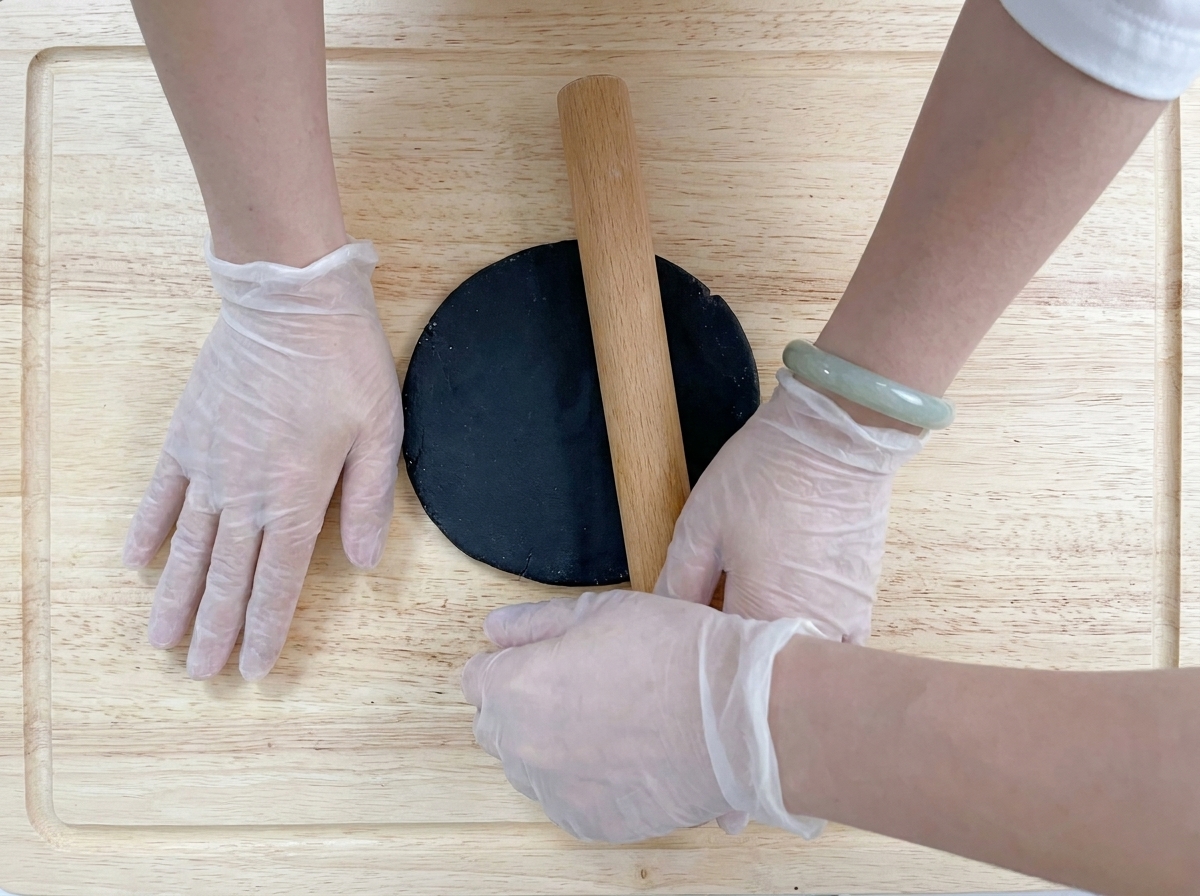 Manos con guantes estirando la masa negra en un círculo plano con un rodillo de madera.