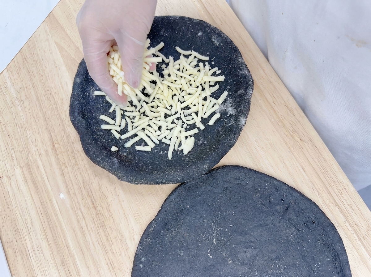 Una mano espolvoreando mozzarella rallada uniformemente sobre la base de pizza negra.