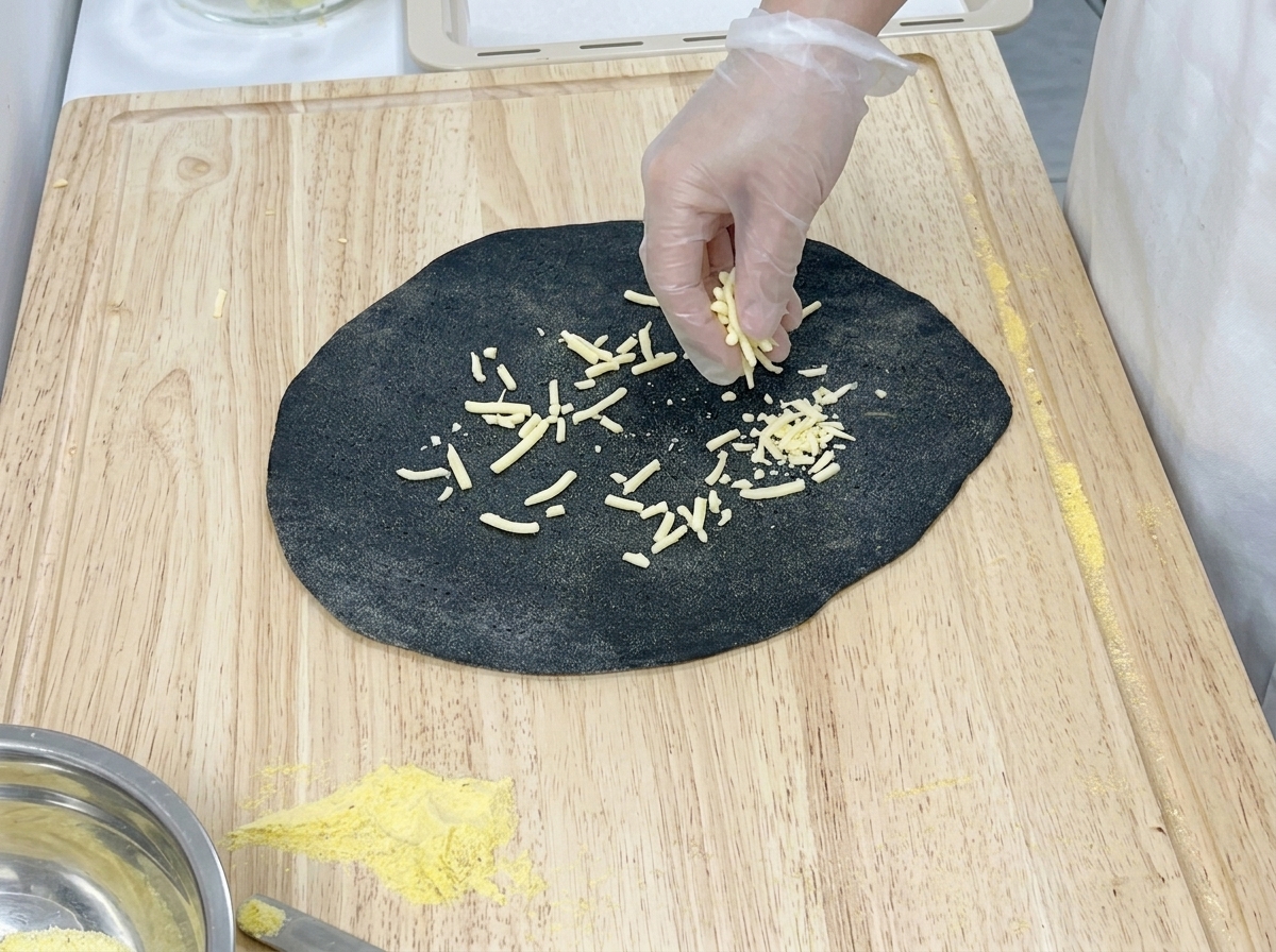 Mano con guante poniendo queso en la mitad derecha de una masa ovalada negra sobre una tabla de madera.