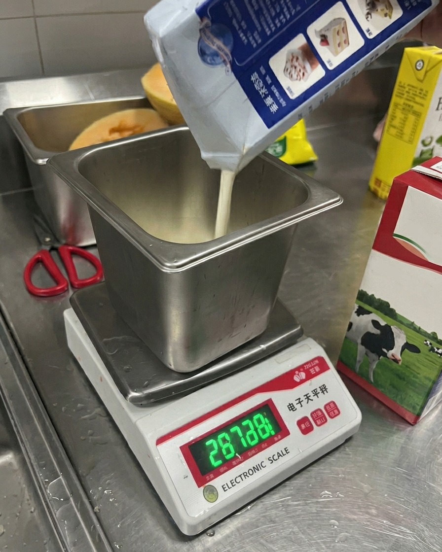 Leche vertiéndose desde un cartón a un recipiente de metal cuadrado sobre una báscula electrónica digital.