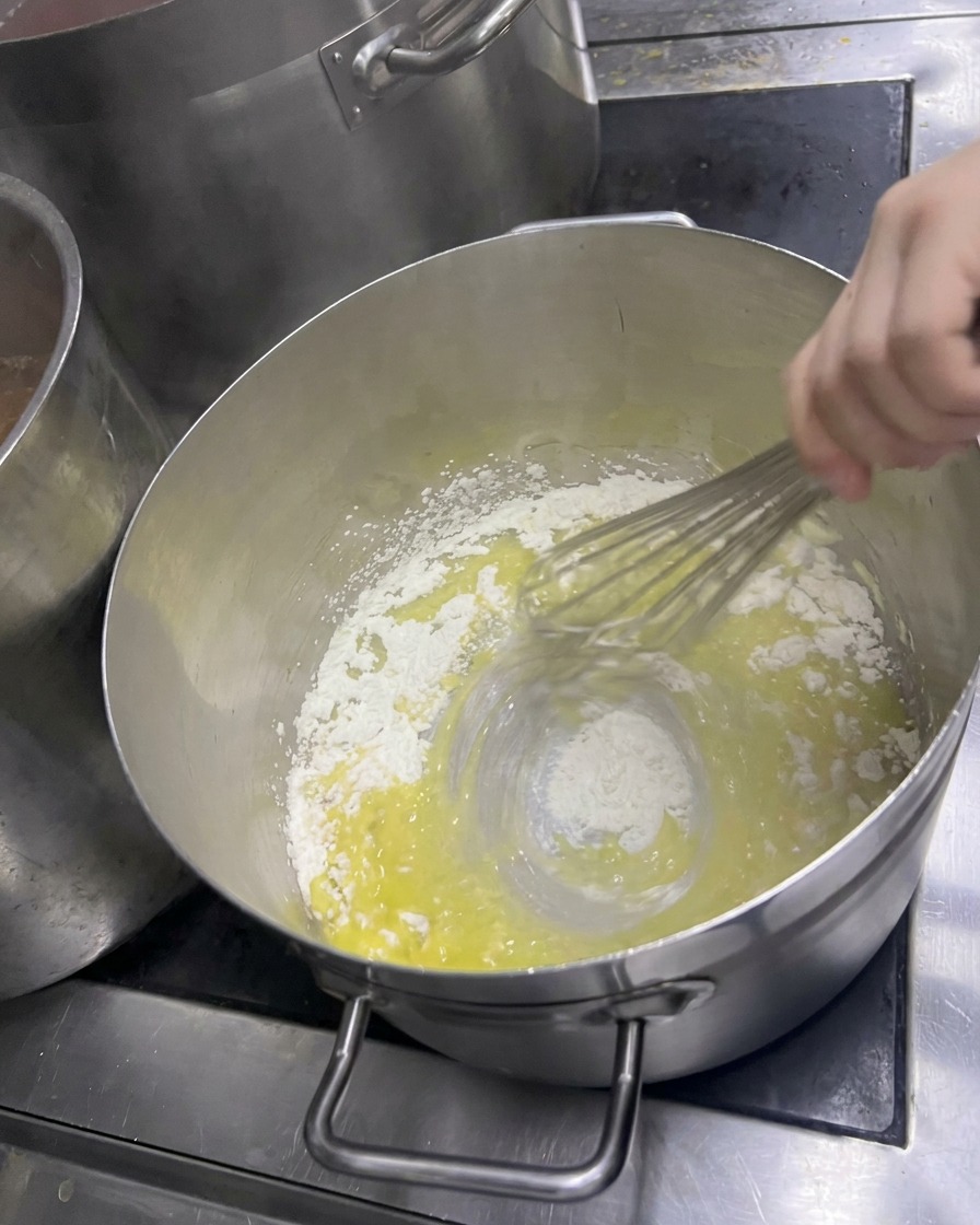 Una mano batiendo vigorosamente harina en mantequilla derretida en una olla para crear un roux amarillo pálido.