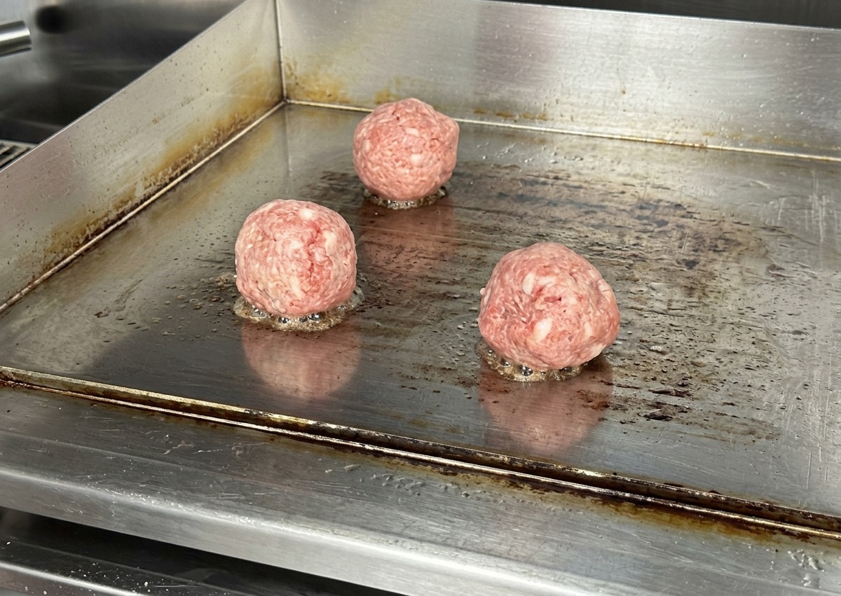 Tres bolas de carne de res cruda sobre una plancha de acero inoxidable caliente con un ligero siseo en la base.