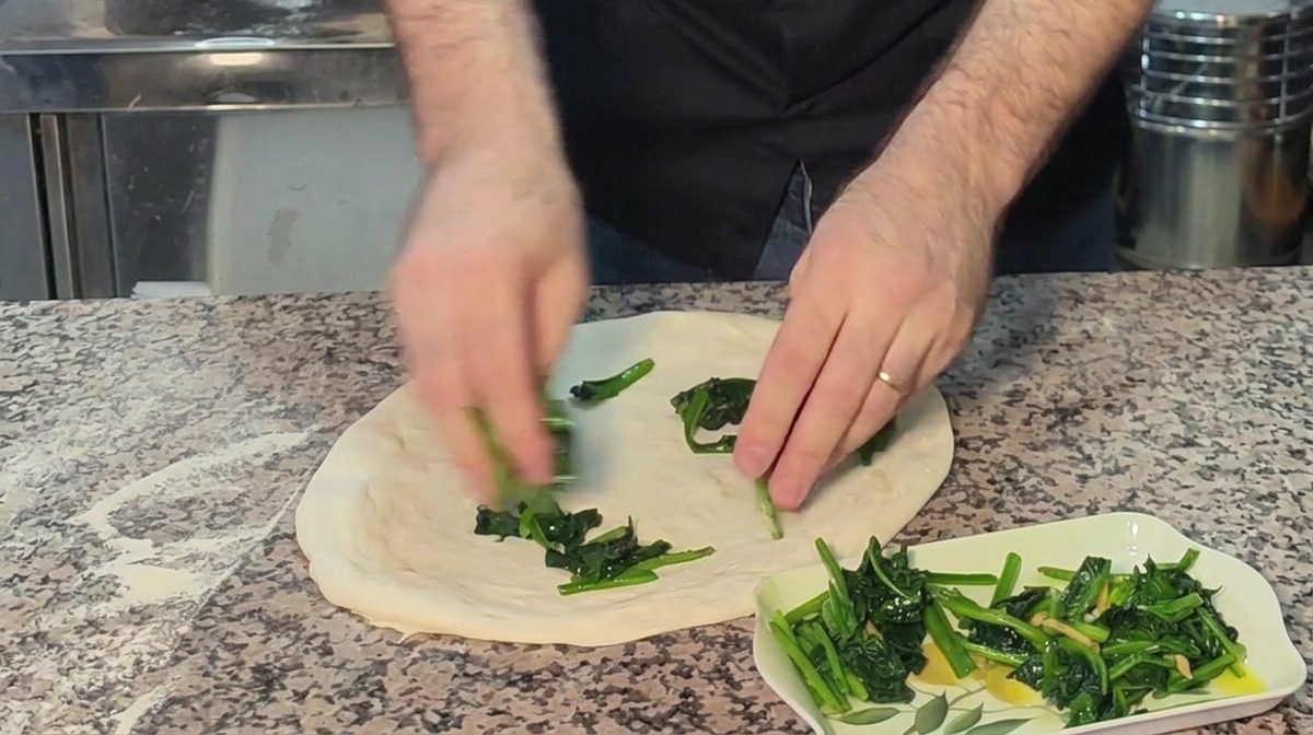 Manos distribuyendo espinacas cocinadas sobre la base de masa de pizza cruda.