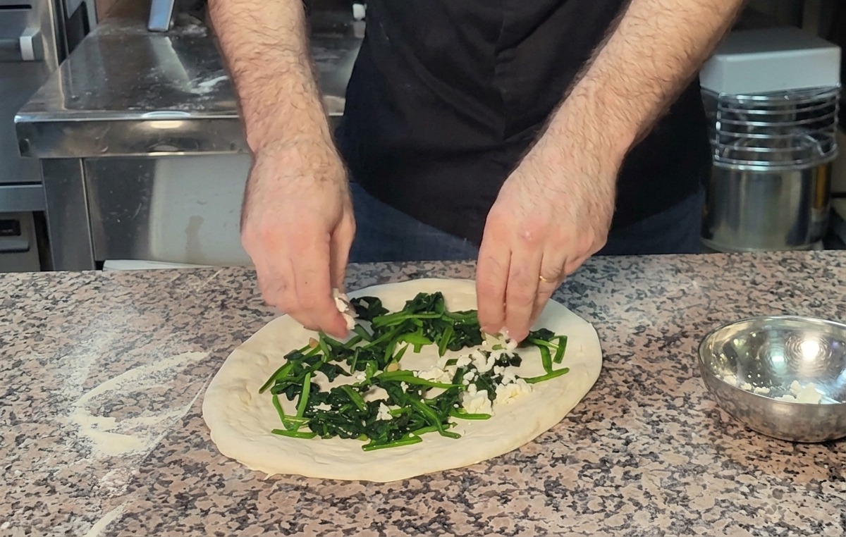 Manos espolvoreando queso feta desmenuzado sobre la capa de espinacas en la masa.