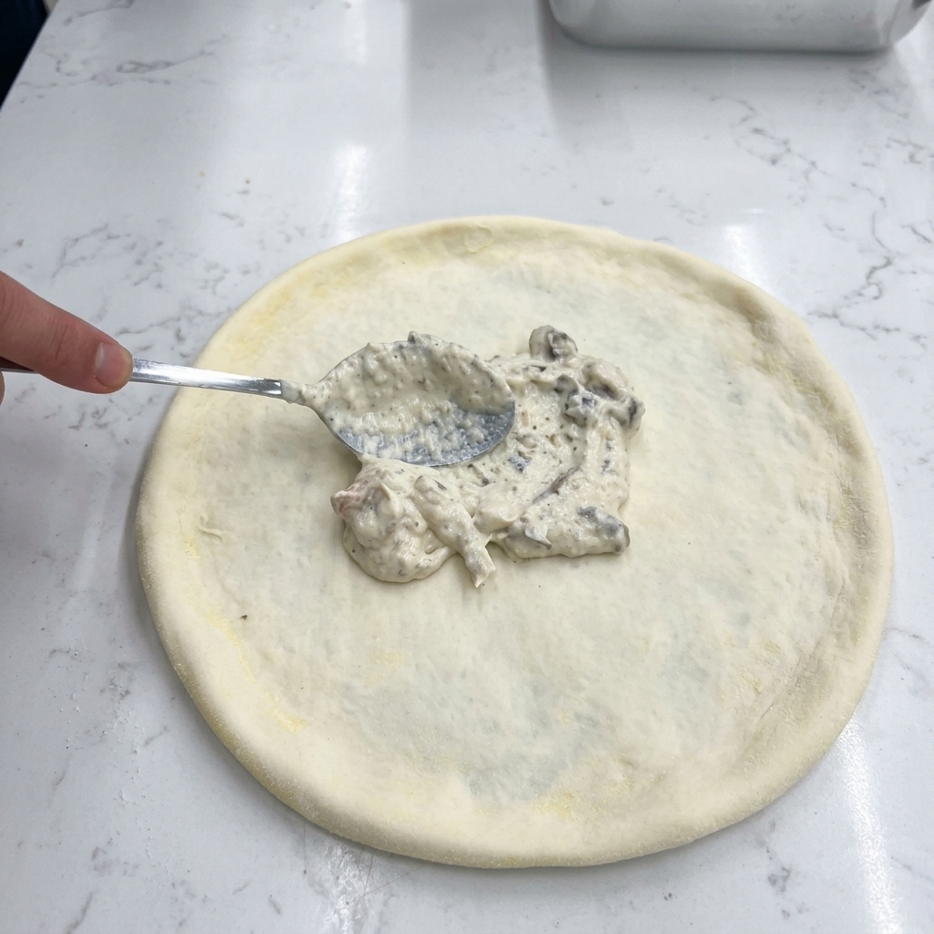 Una mano usando una cuchara metálica para extender salsa cremosa de champiñones sobre la masa cruda.