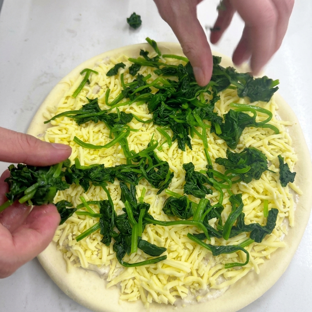 Dos manos colocando trozos de espinacas blanqueadas sobre la base con queso mozzarella.