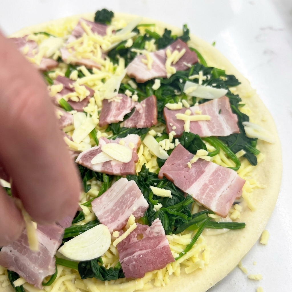 Una mano esparciendo queso mozzarella sobre la pizza cruda con espinacas, beicon y ajo.