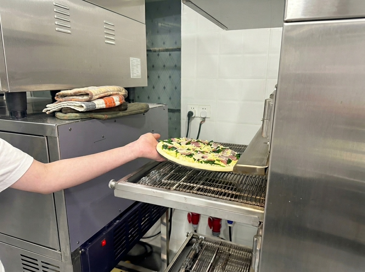 Un brazo colocando la pizza de espinacas y beicon en la cinta transportadora de un horno de pizza comercial.